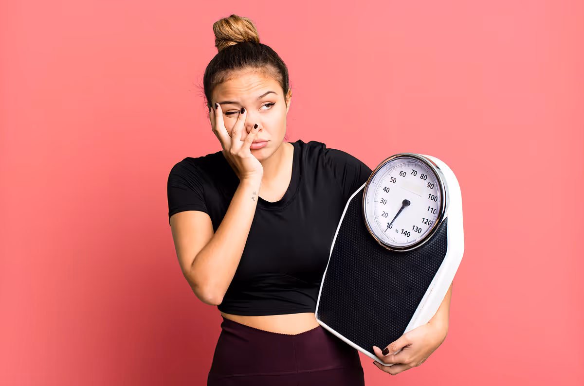Tired young woman holding a weight scale and rubbing her eye against a pink background.