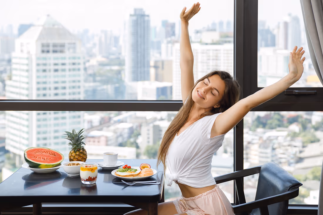 Young woman stretching with eyes closed at a table with a breakfast spread including watermelon, pineapple, yogurt, coffee, and toast in front of a large window showing a cityscape.
